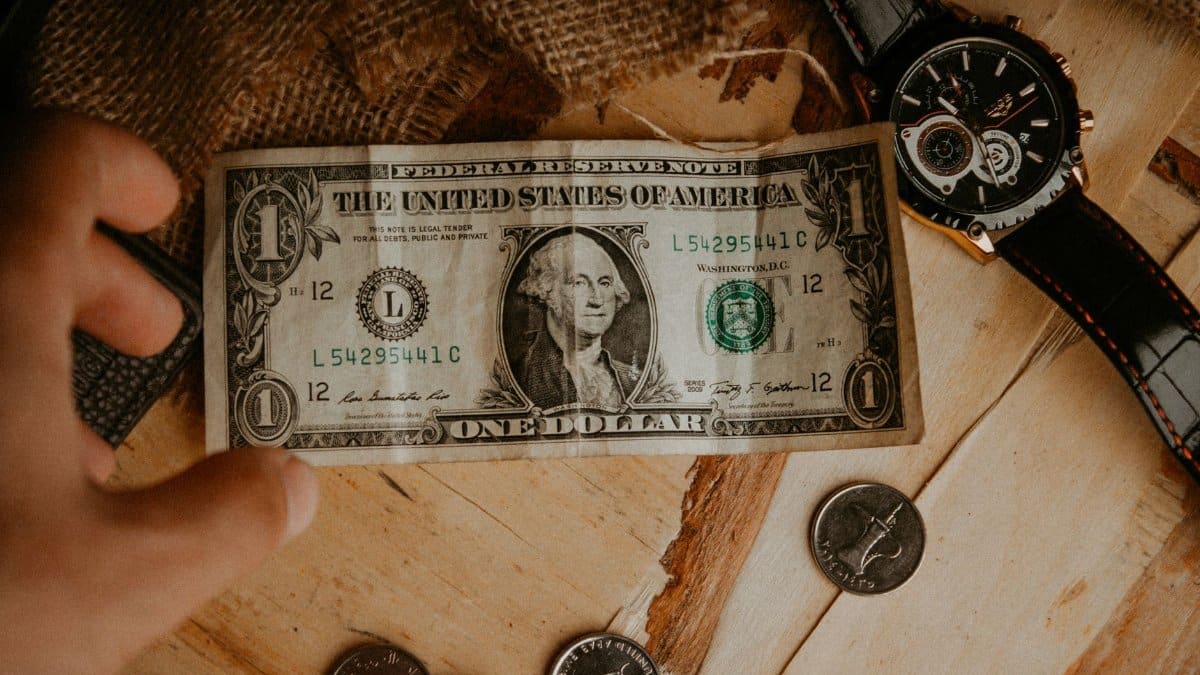 A person reaches for a one dollar bill surrounded by coins and a watch on a wooden table.