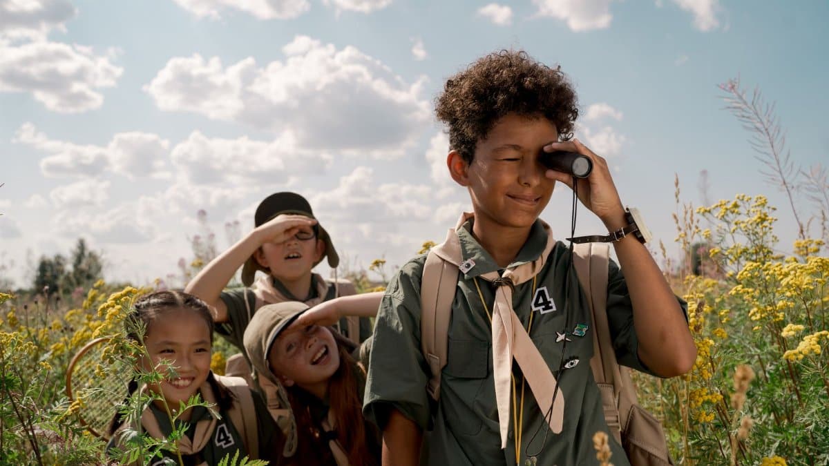 A group of kids in scout uniforms exploring nature with a spotting scope on a sunny summer day.