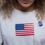 A woman with curly hair holding an American flag and wearing a vote pin against a blue background.