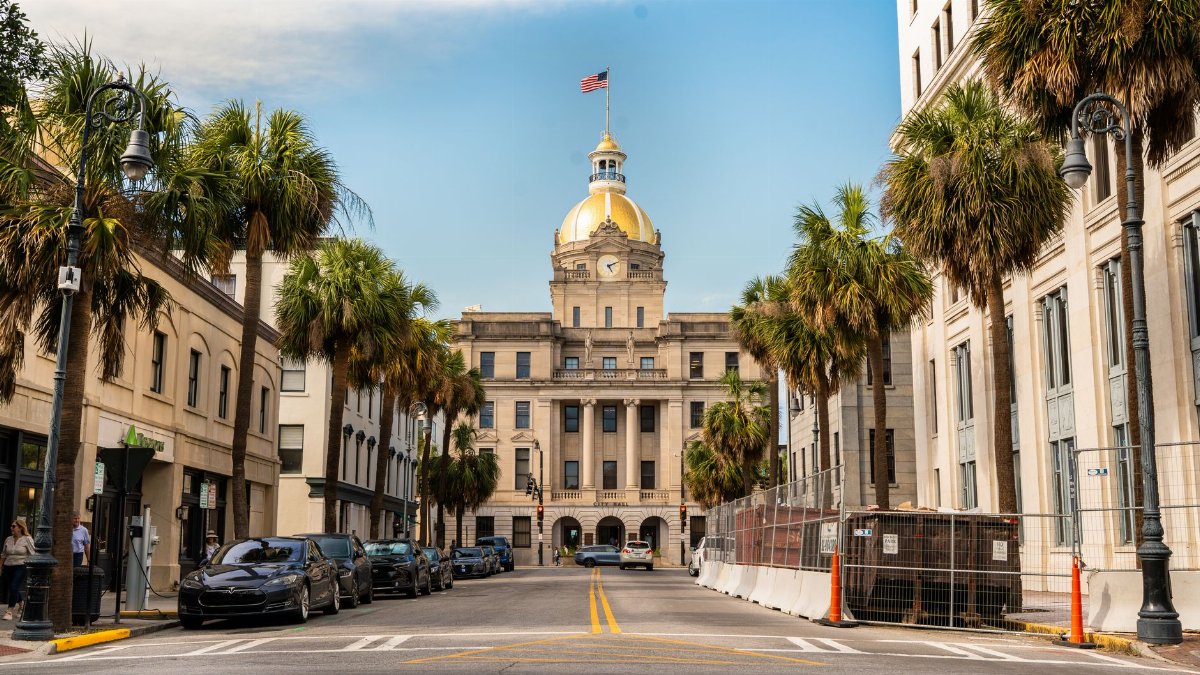 View of Savannah City Hall with its golden dome framed by palm trees along a downtown street.
