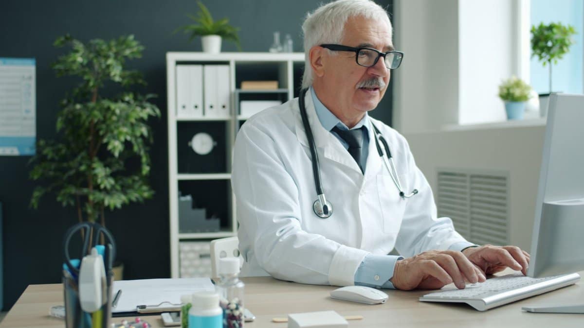 Elderly male doctor typing on keyboard in modern medical office.