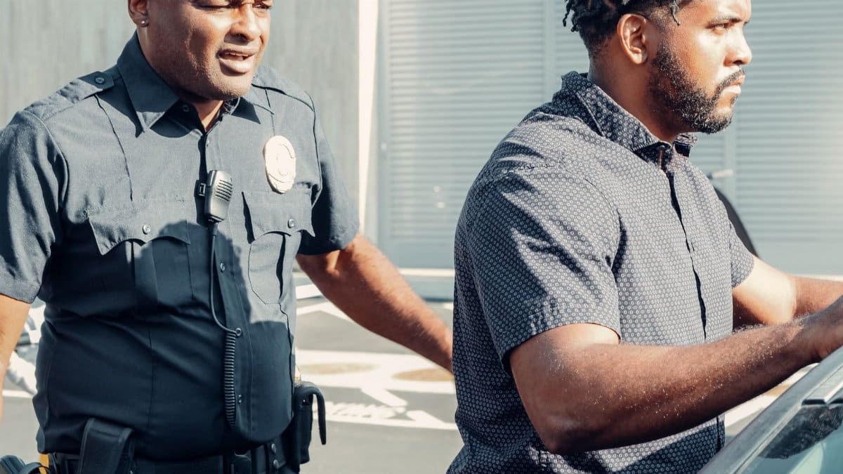 A police officer detaining a man beside a car in an outdoor setting.