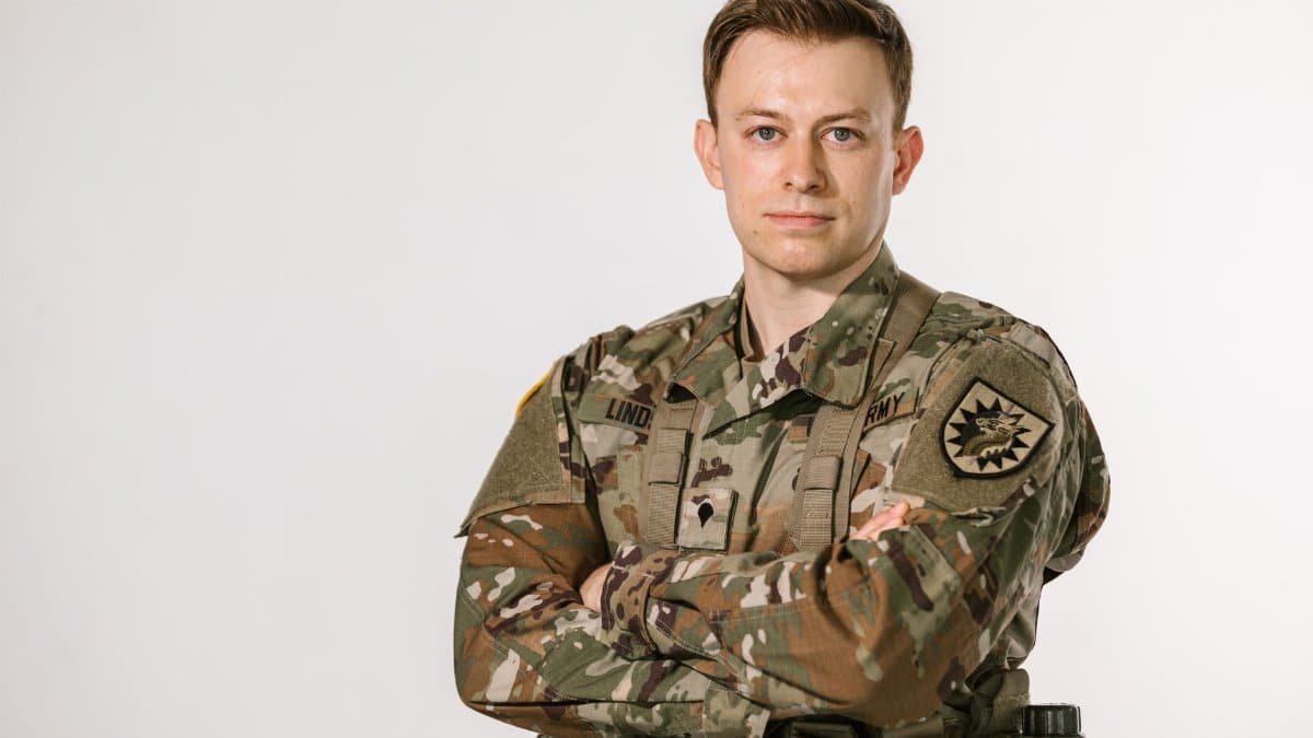 A proud US Army soldier in camouflage uniform standing with crossed arms in a studio setting.