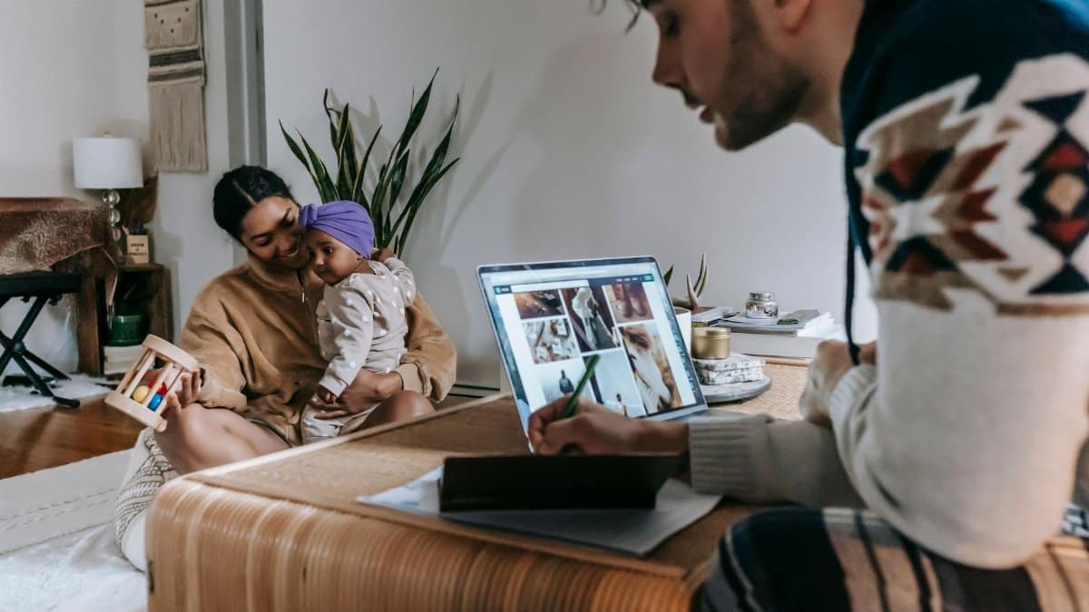 Crop pensive ethnic father writing in notebook and working with laptop while ethnic mother playing with cute little daughter