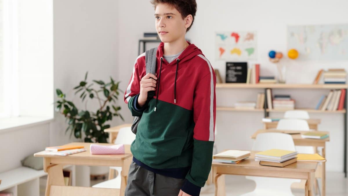 Teen boy with backpack in a colorful hoodie standing in a classroom setting.