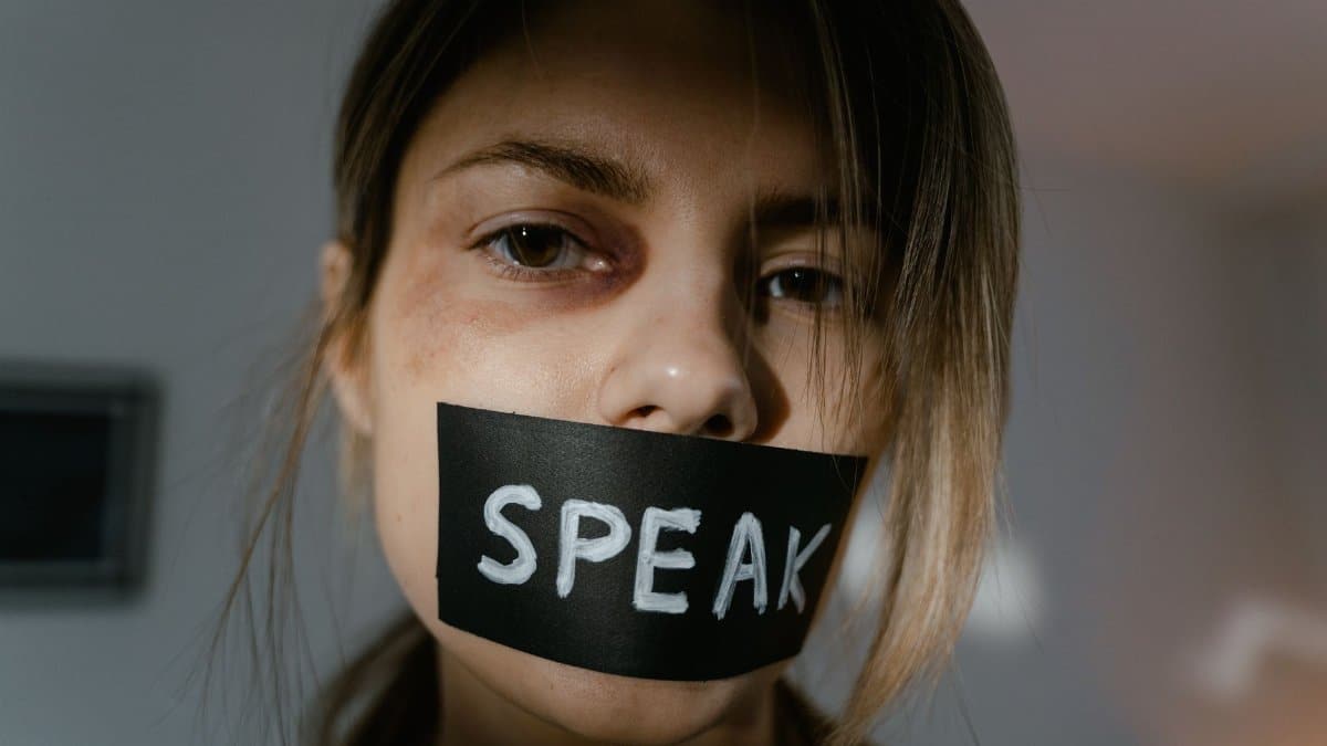 Close-up of a woman with 'Speak' tape across her mouth, conveying silence.