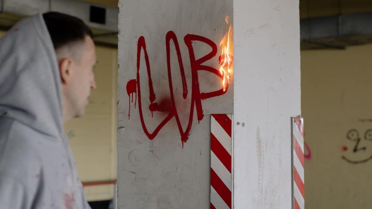 Man in hoodie observes burning graffiti on wall in urban location, showcasing street art and fire.