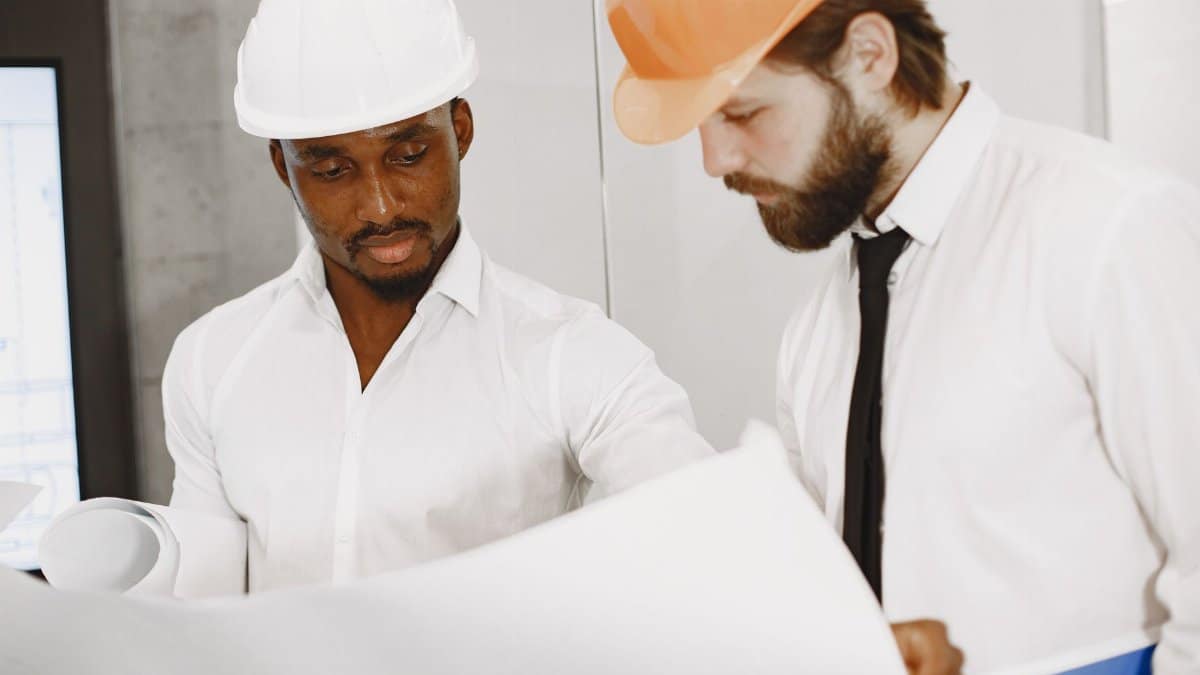 Two engineers in hard hats concentrate on reviewing detailed construction blueprints indoors.