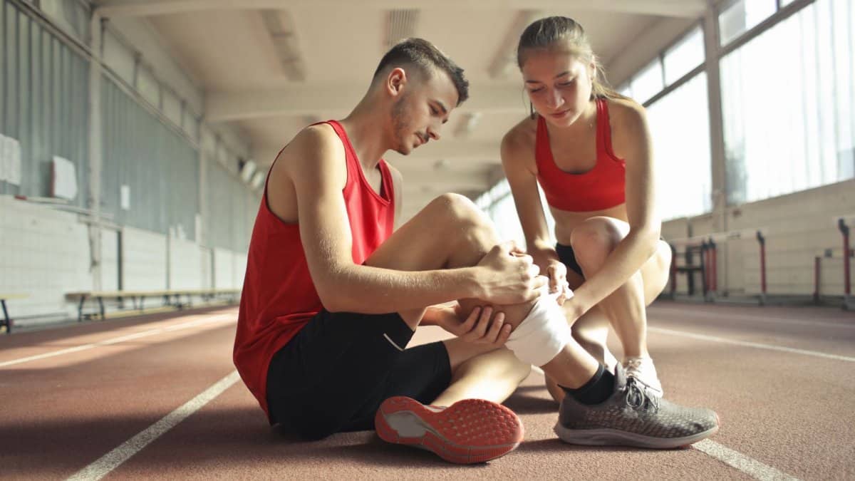 Side view of athlete sitting on rubber track in sports hall while sportive female partner helping to band leg after injury