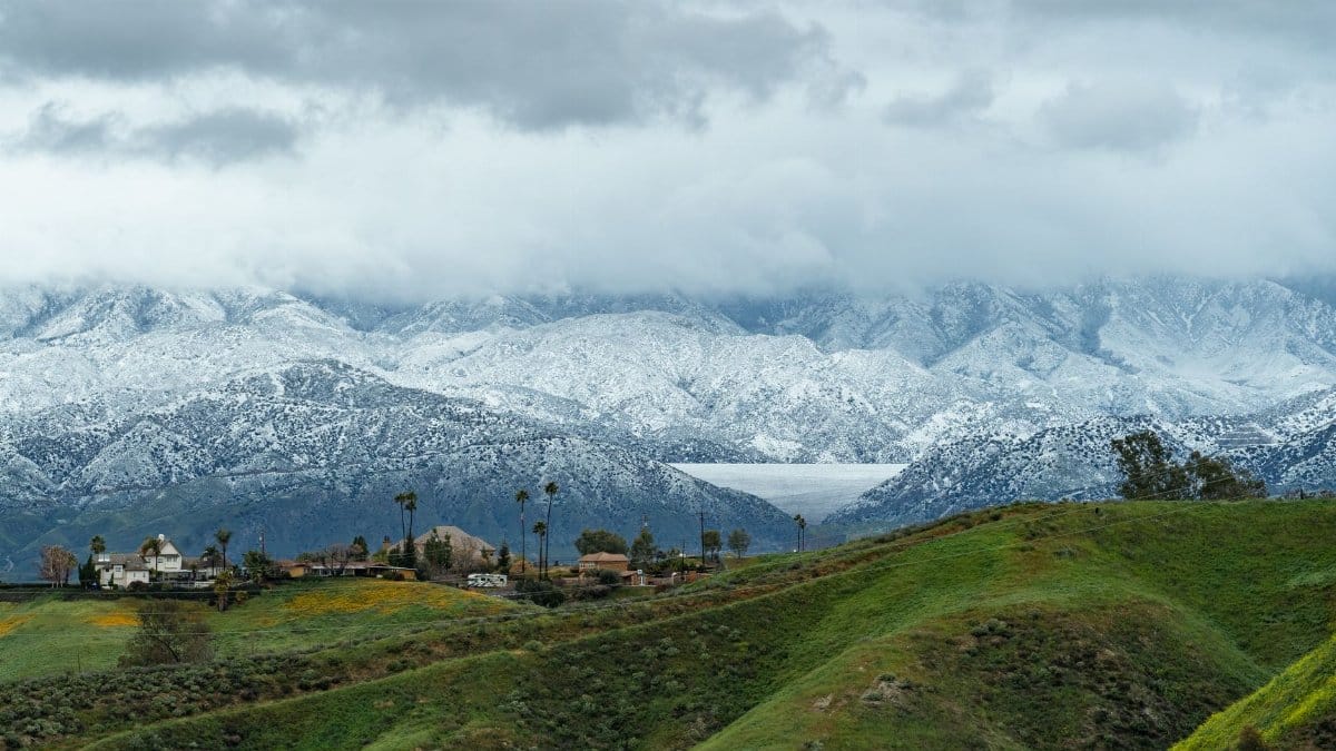 Picturesque view of snow-covered San Bernardino Mountains with green hills and cloudy sky in Redlands, CA.