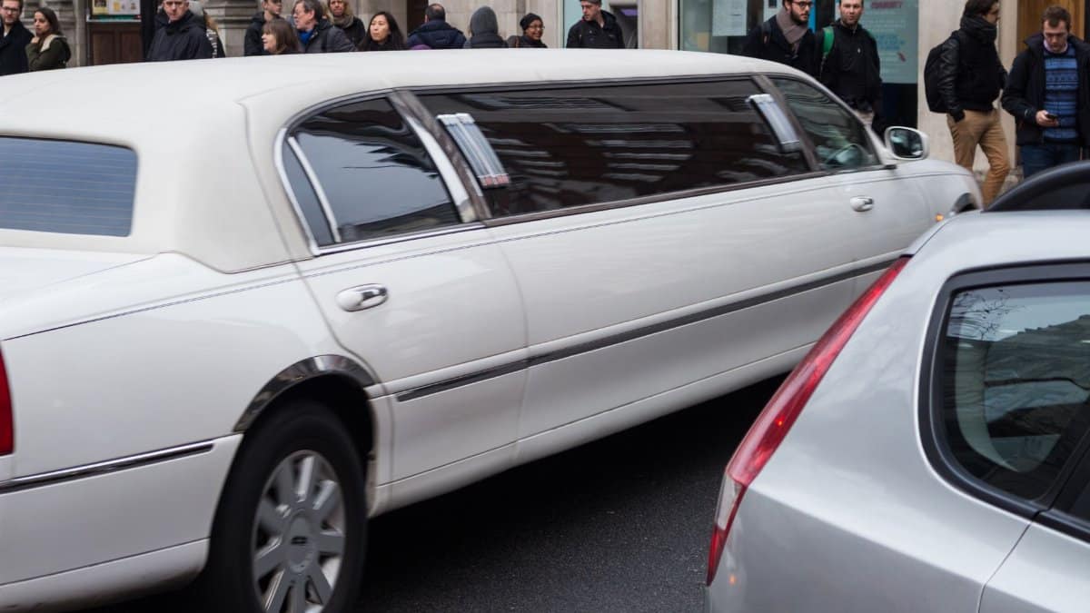 A white limousine driving down a bustling city street with pedestrians and other vehicles.