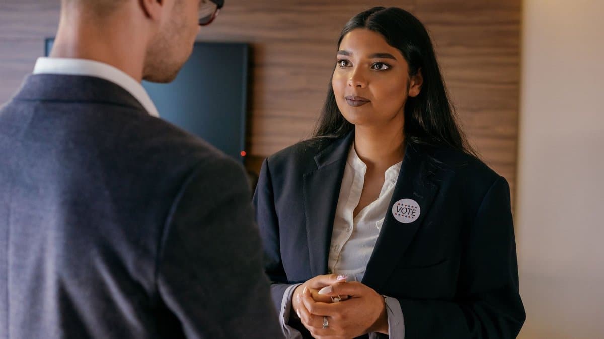 A female candidate in a political discussion, wearing a vote badge during an election campaign indoors.