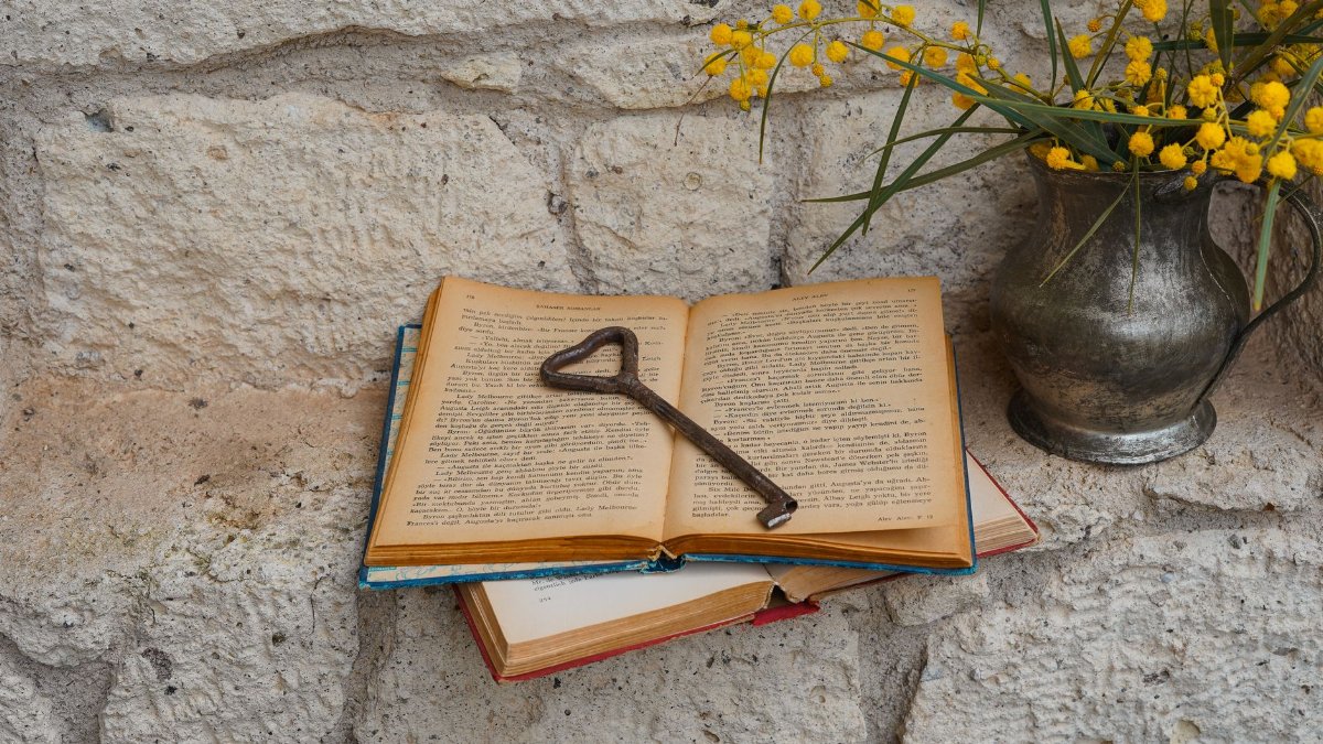 Vintage open book with antique key, silver vase, and yellow mimosa on stone surface.