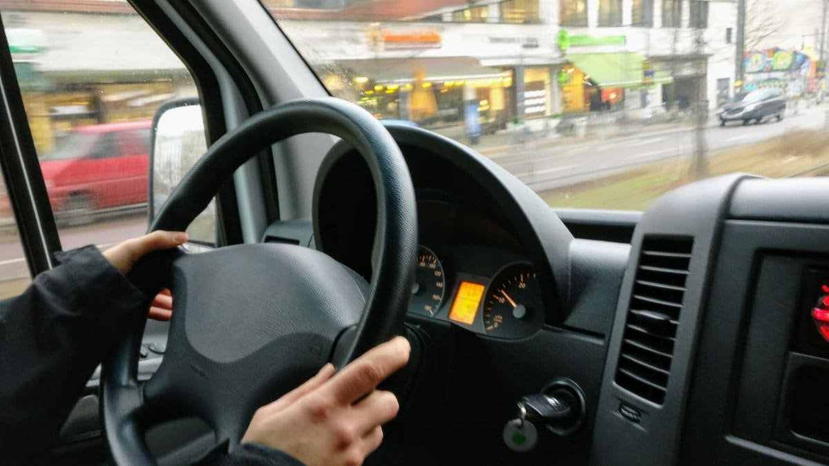 Hands on a steering wheel driving a van through an urban area with shops visible outside.