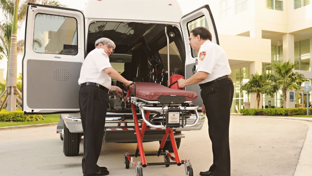 Two paramedics load a stretcher into an ambulance on a sunny day outside a modern hospital.