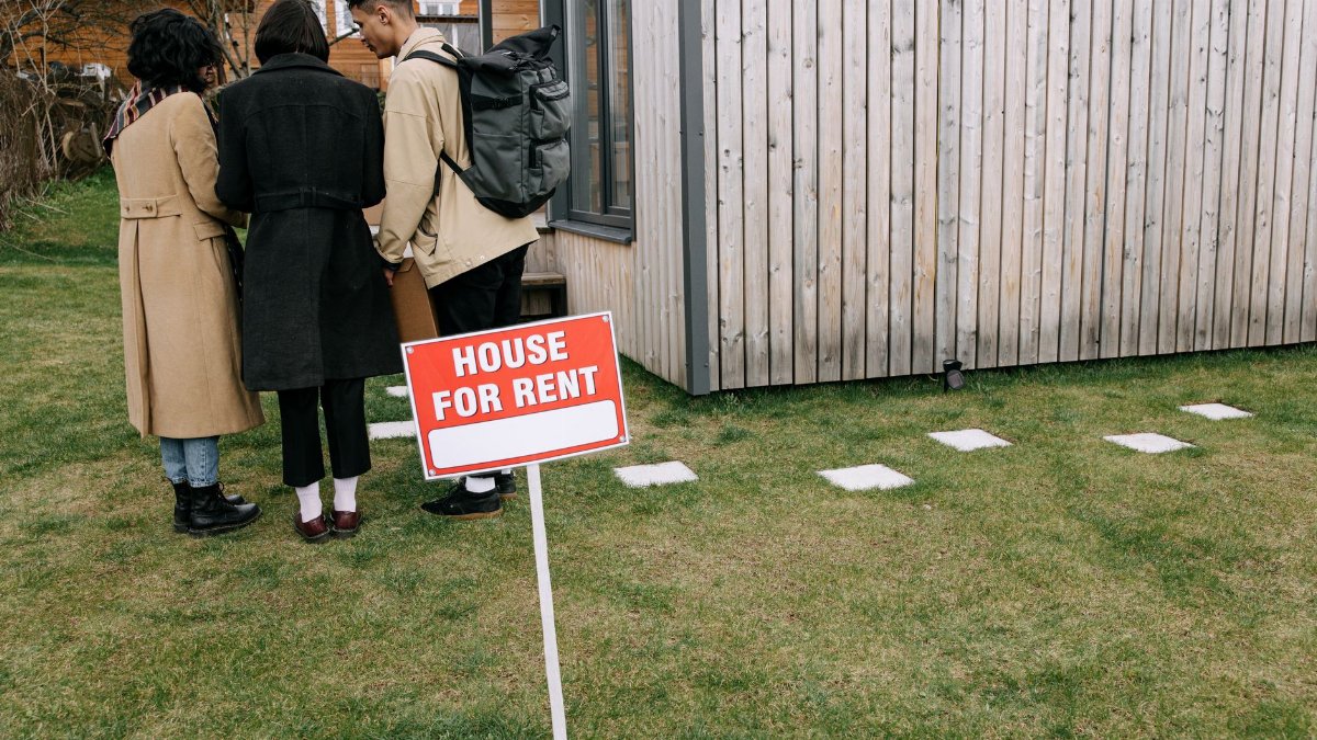 Group of adults viewing a wooden house with a 'House for Rent' sign on the lawn.