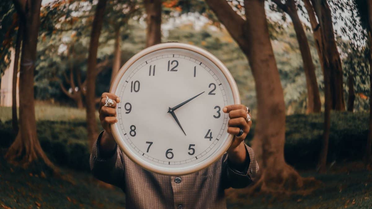 An individual holding a large clock in an outdoor forest environment, symbolizing time.