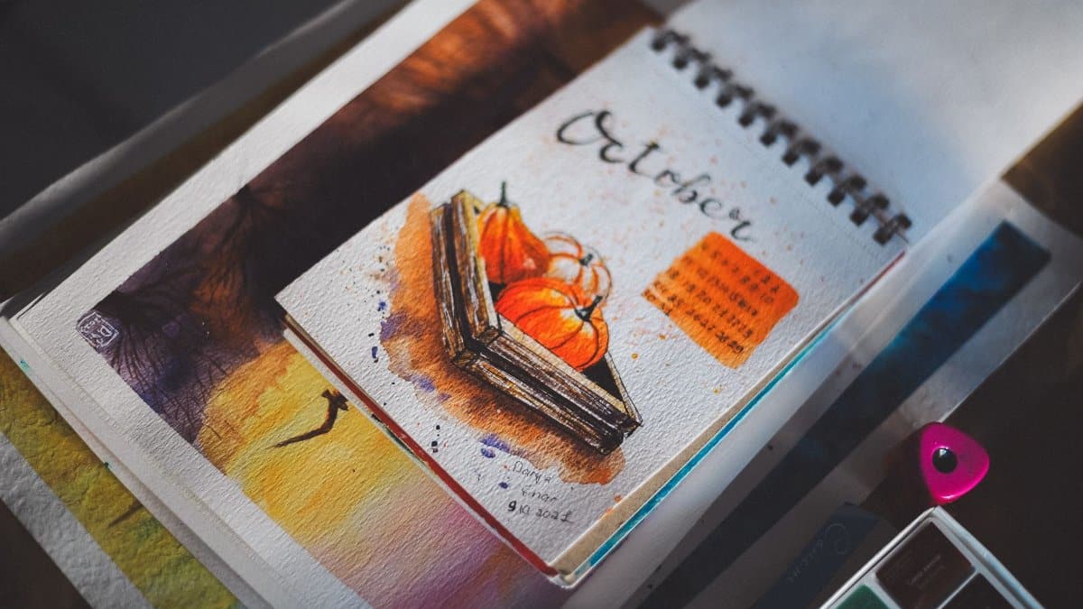 Open sketchbook showing a vibrant October watercolor painting with pumpkins and a calendar.