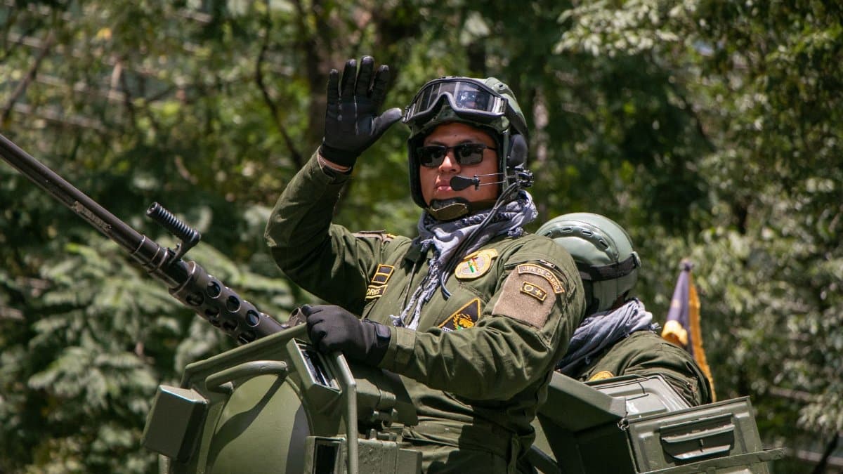 Military personnel wave from an armored vehicle in a lush forest setting.