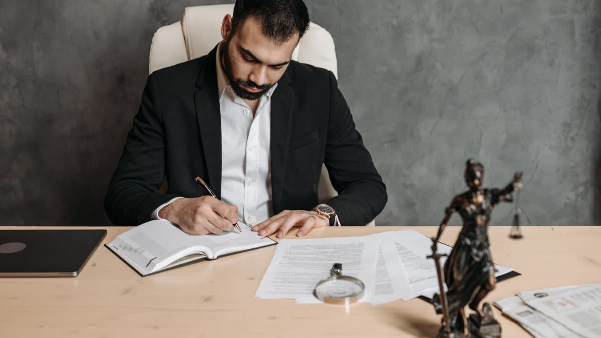 Focused lawyer in black suit at desk writing on documents in an office setting with legal statue.