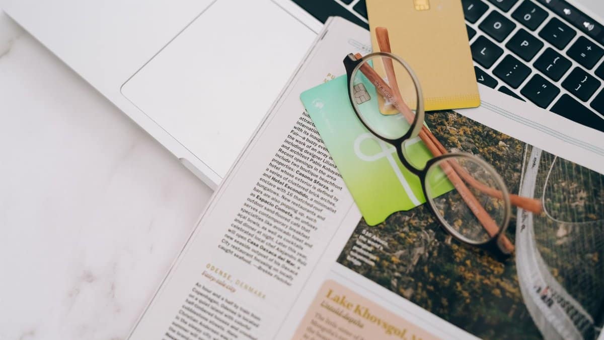Eyeglasses resting on a magazine beside a laptop and credit cards, indoors.
