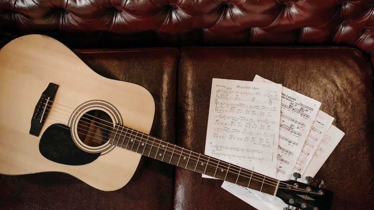 A flat lay of an acoustic guitar with sheet music on a leather sofa, perfect for music lovers.