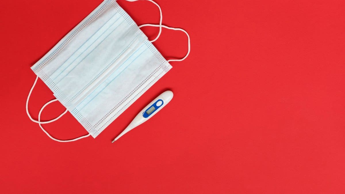 Flat lay of a surgical mask and digital thermometer on a red background, symbolizing health and safety.