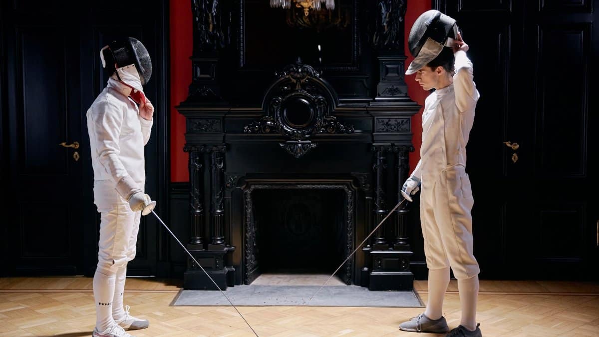 Two fencers preparing for a duel in a stylish indoor setting with a fireplace.
