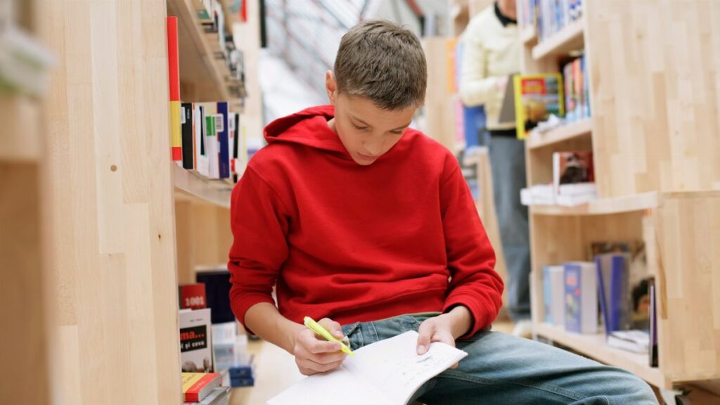 A teenager in a red sweater studying in a library, surrounded by bookshelves.