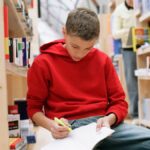 A teenager in a red sweater studying in a library, surrounded by bookshelves.