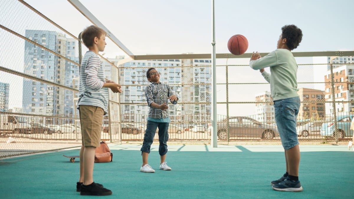 Three boys playing basketball on an outdoor court surrounded by city buildings.