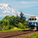 Passenger train traveling with Mount Rainier in the background, lush greenery, bright skies.