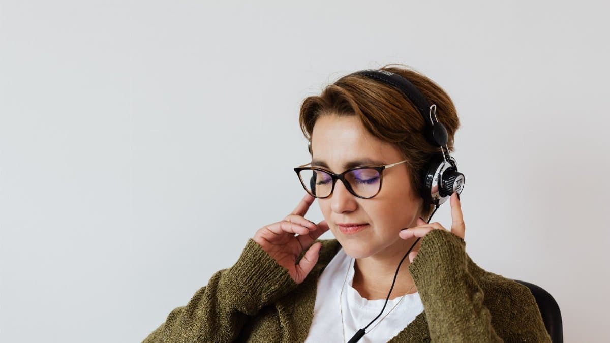 Content glad female wearing eyeglasses and headphones listening to good music and touching headset while sitting with eyes closed against white wall