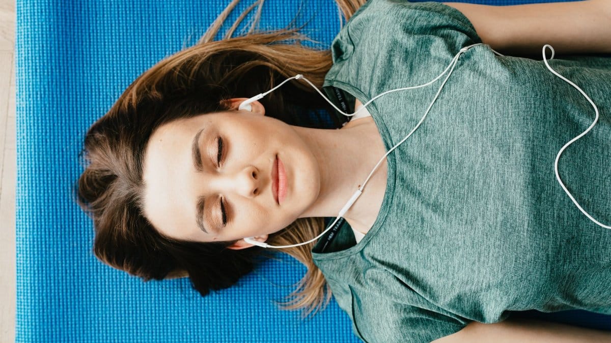 From above of calm young female in casual wear listening to music in earphones while taking break after workout on blue mat in flat