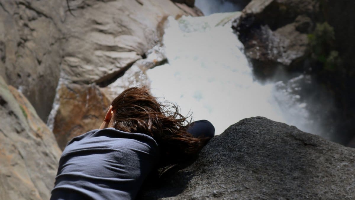 A woman adventurer peers over a cliff at a cascading waterfall, exemplifying thrill and nature's power.