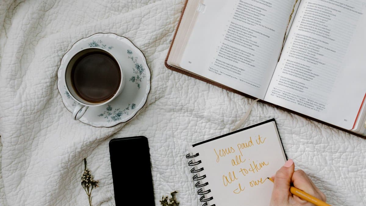 Flat lay of Bible study with coffee, notebook, and phone on a quilted blanket.