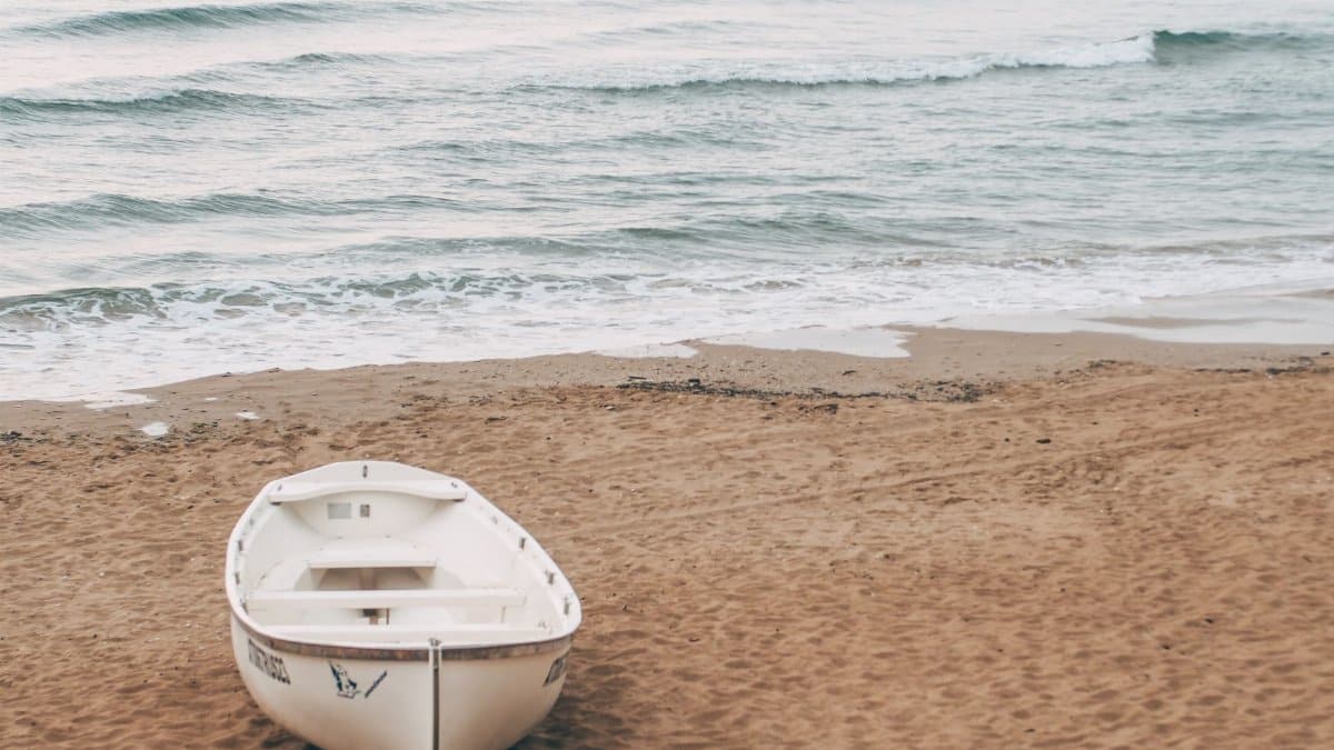 A white boat rests on a sandy beach, with gentle waves lapping the shore under a calm sky.
