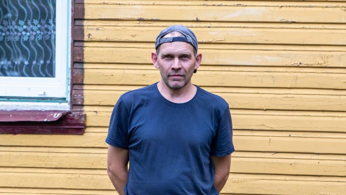 Portrait of a rural Estonian man standing in front of a traditional wooden house in Tartu County.