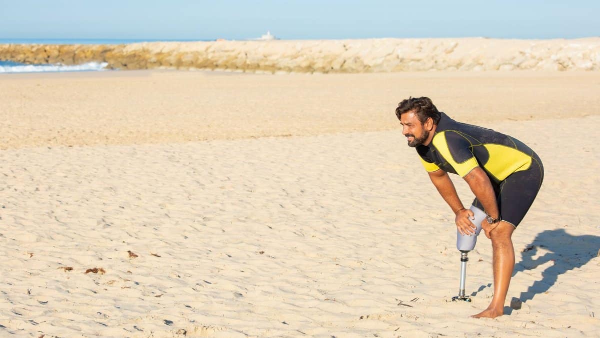 Side view full length strong tired male athlete with leg prosthesis standing in recovery position with hands on knees during intense workout on sandy seashore