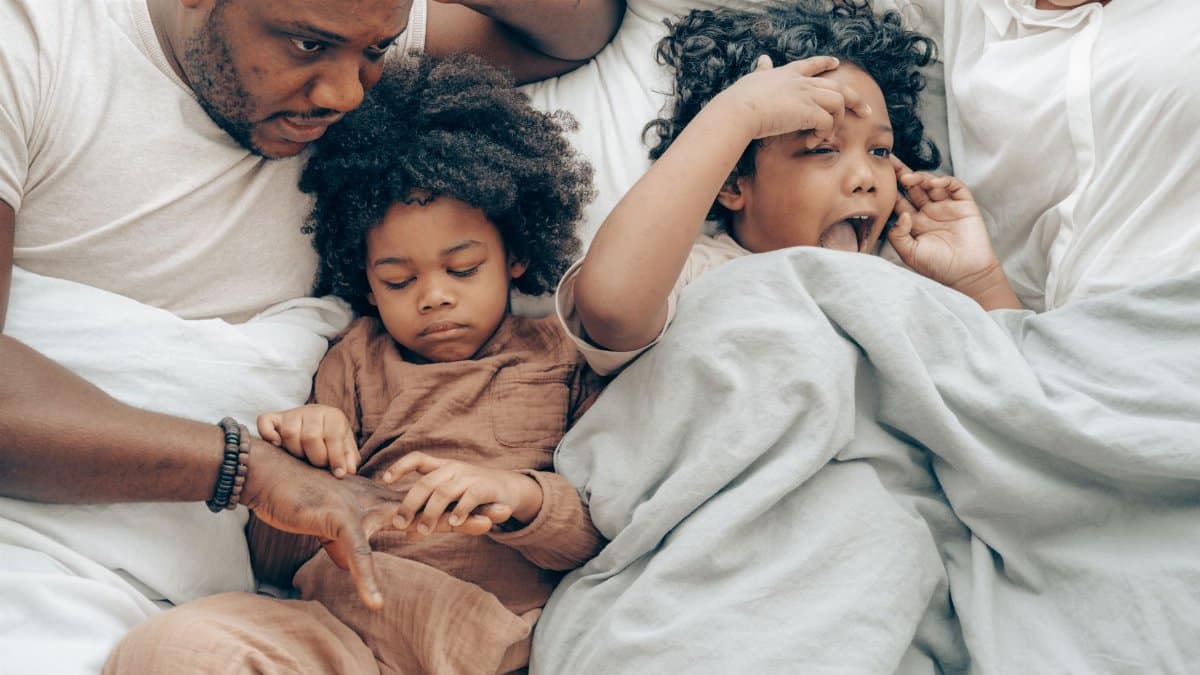 From above of happy little ethnic girls cuddling crop parents while lying together in bed in morning