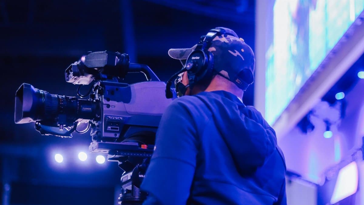 A dedicated cameraman wearing a headset and cap operates a large professional camera at an indoor live event.