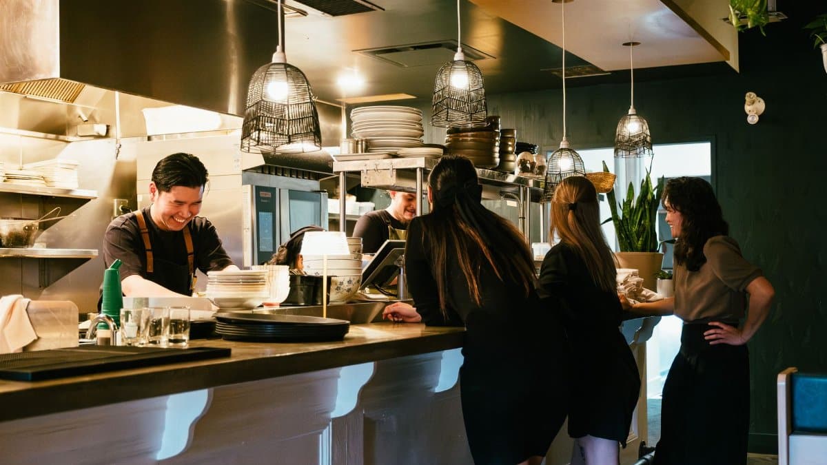 Busy restaurant kitchen with staff preparing meals as patrons watch. Warm lighting and informal dining atmosphere.