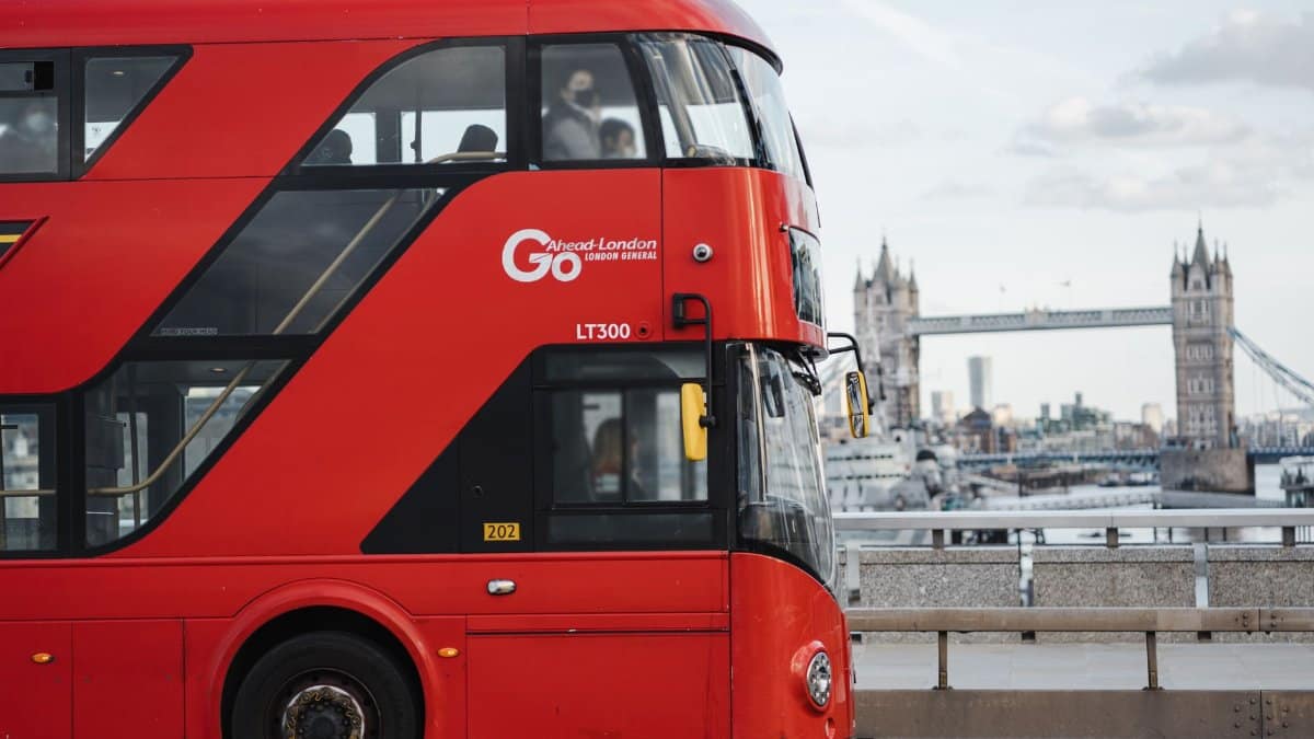 Contemporary double decker red bus driving on road along calm river against historic Tower bridge and residential buildings in London city
