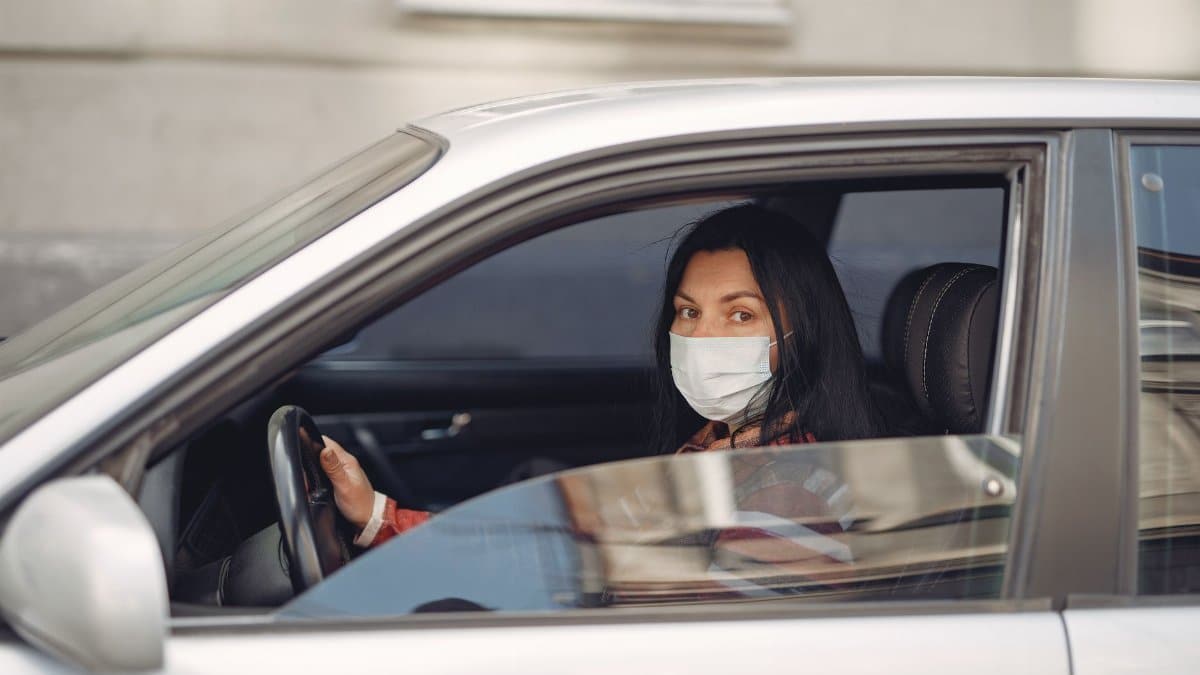 Female driver wearing casual outfit and protective mask on face sitting in automobile with opened window during Coronavirus pandemic near city building and looking at camera