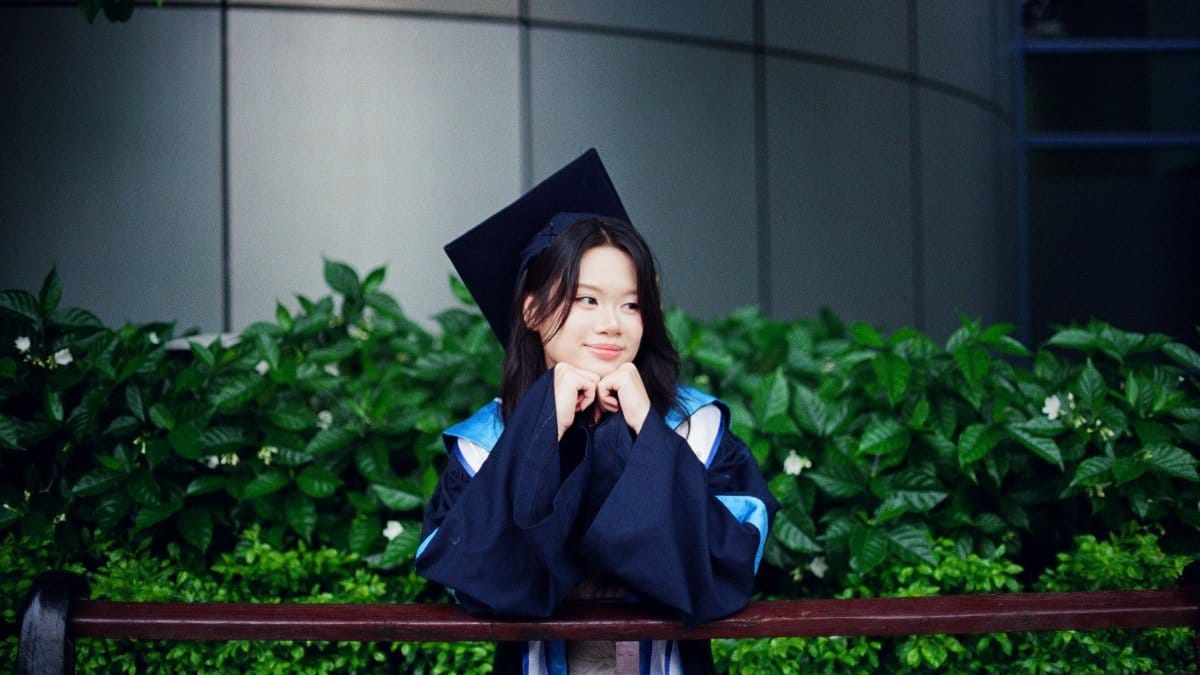 A young graduate in a cap and gown sitting on a bench outdoors with greenery.