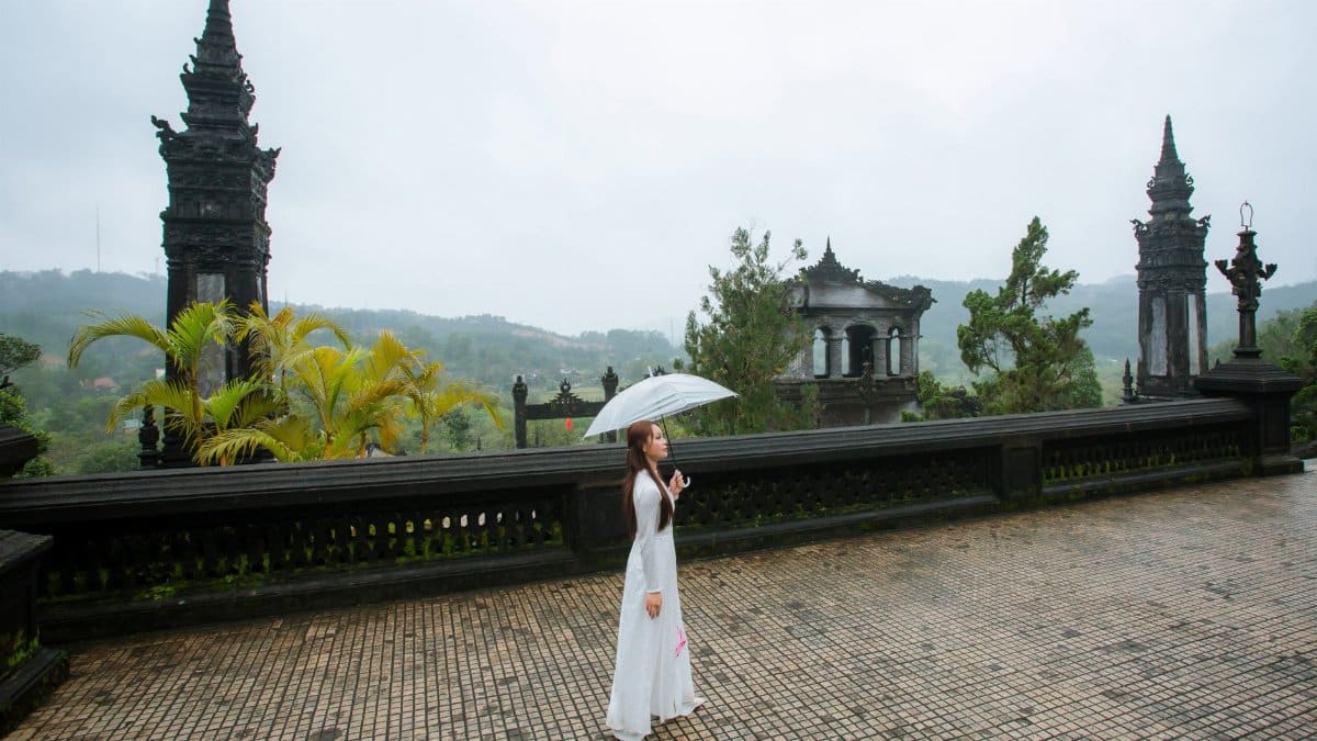 Woman in a traditional long dress with an umbrella at a temple in Vietnam, showcasing cultural elegance.