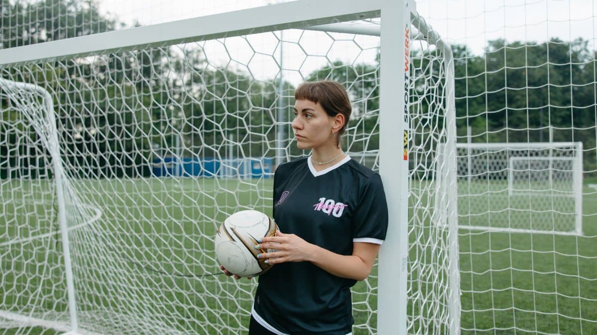 Confident female soccer player holding ball on a green field by the goalpost.