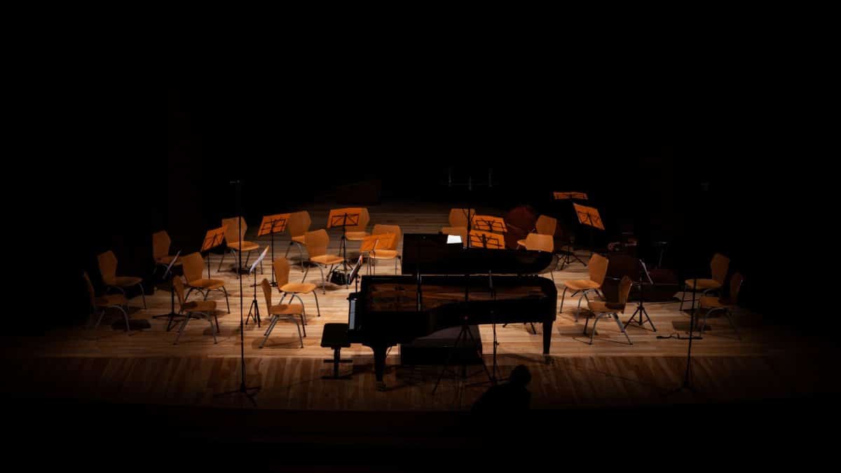 A dimly lit concert hall stage featuring an empty grand piano and chairs for an orchestra.