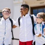 Three smiling schoolchildren with backpacks outside a school building, enjoying their first day back to school.