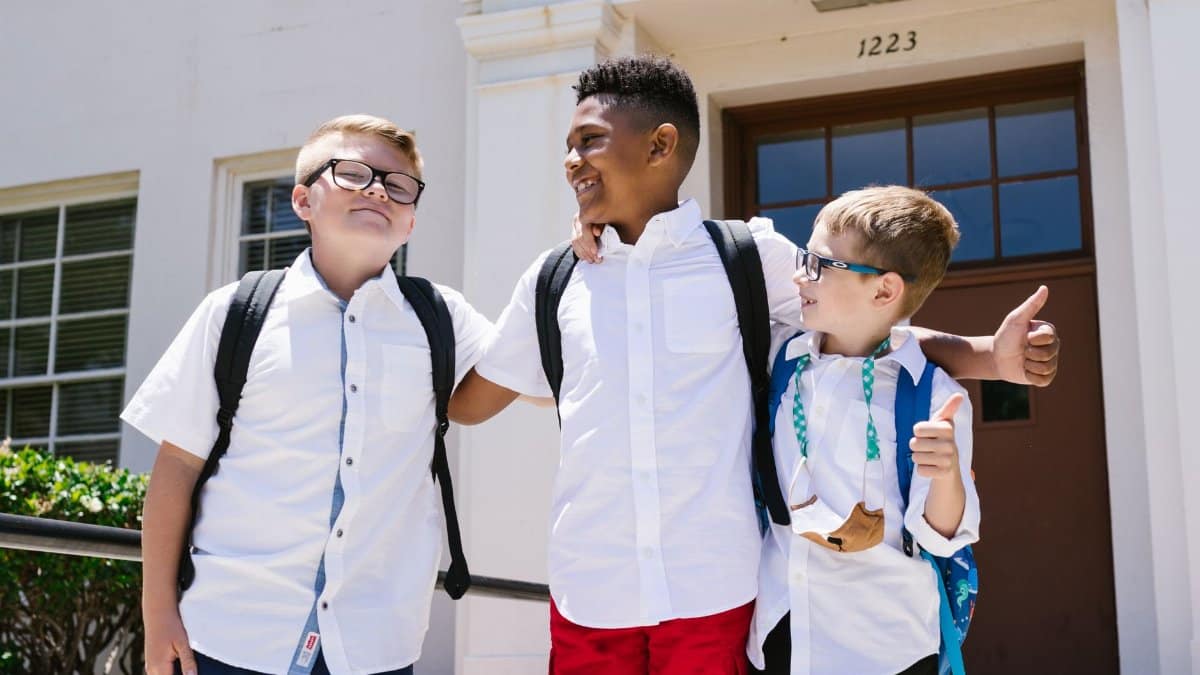 Three smiling schoolchildren with backpacks outside a school building, enjoying their first day back to school.